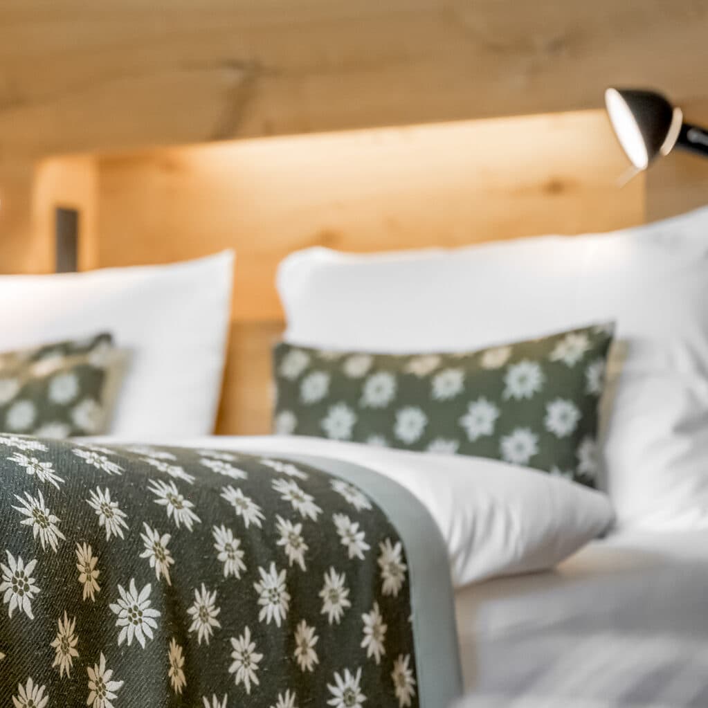 Close-up of a neatly made bed with white pillows and green, floral-patterned pillowcases and a matching bed runner, against a wooden headboard with a reading lamp.