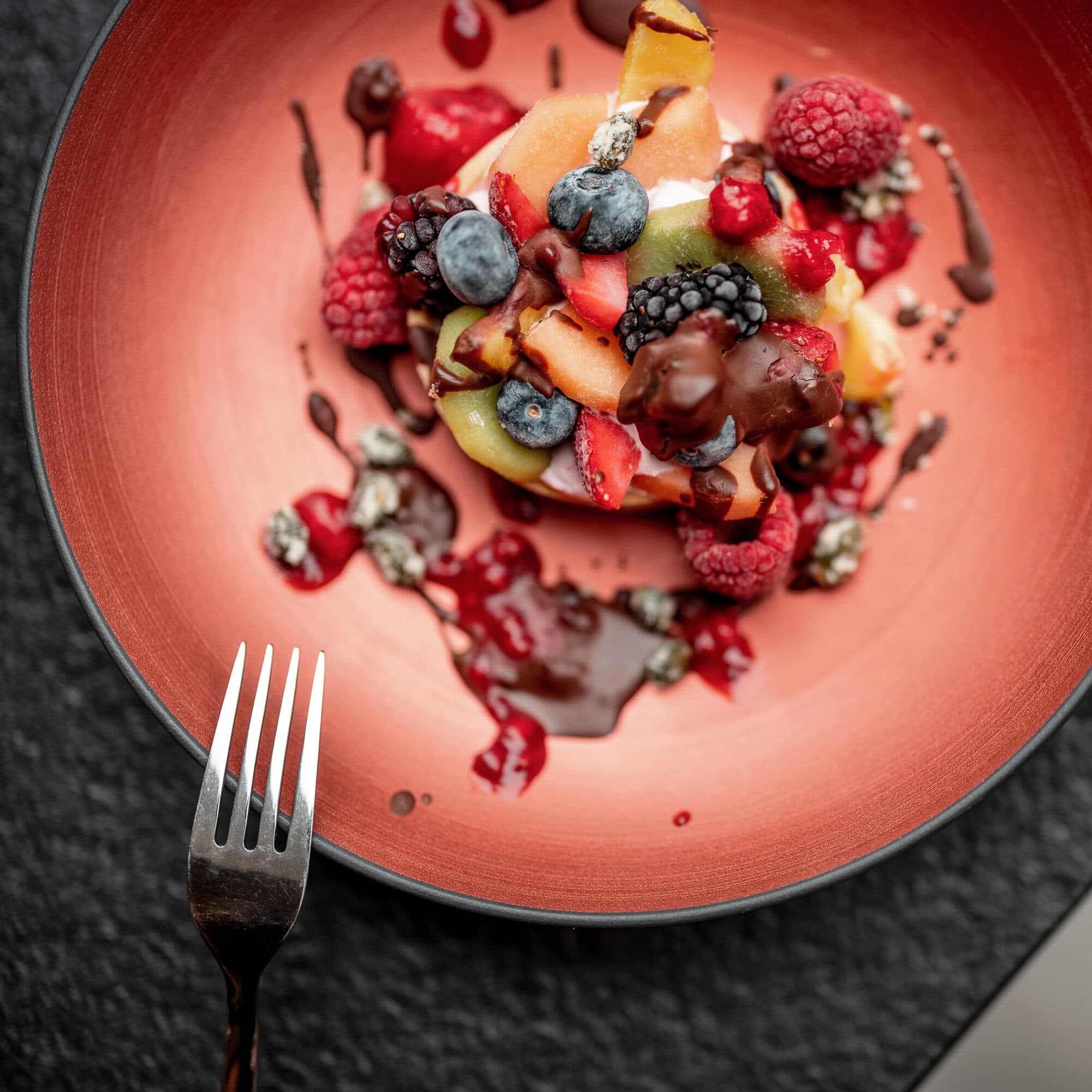 Un piatto da dessert con frutti di bosco freschi assortiti, kiwi e melone, irrorati di salsa al cioccolato, si trova accanto a una forchetta su una superficie scura e strutturata.
