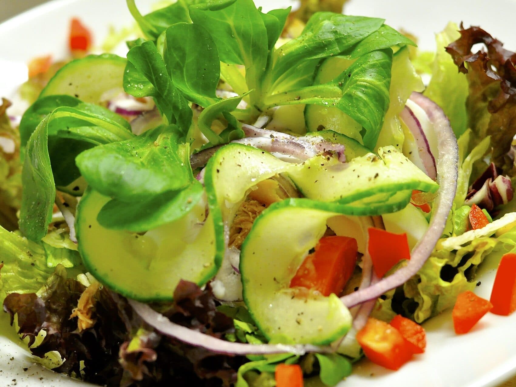 Ein frischer Salat mit gemischtem Grün, geschnittenen Gurken, roten Zwiebeln und gewürfelter roter Paprika, garniert mit schwarzem Pfeffer.