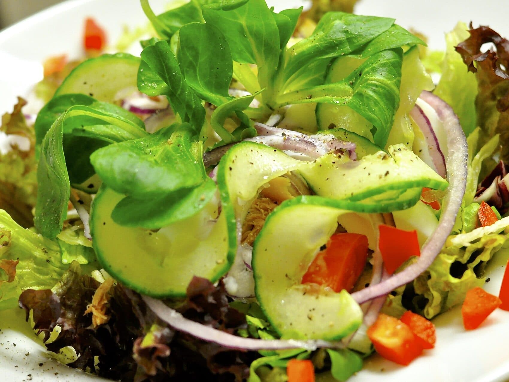 Une salade fraîche avec des feuilles vertes, des tranches de concombre, de l'oignon rouge et des cubes de poivron rouge, assaisonnée de poivre noir.