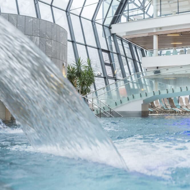Piscina coperta con acqua a cascata, pareti moderne in vetro, piante in vaso e una fila di sedie a sdraio sullo sfondo.