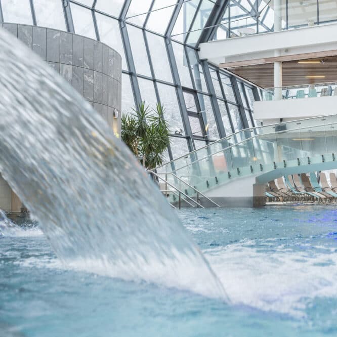 Piscine intérieure avec une cascade incurvée, des murs en verre, un escalier moderne et des chaises longues disposées le long de la piscine.