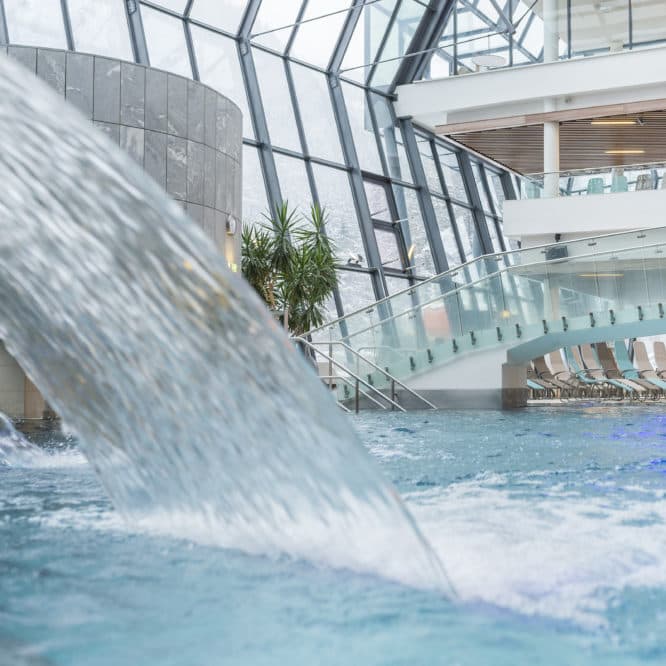 Innenpool mit Wasserspiel, Glaswänden, geschwungener Treppe, Topfpflanzen und Sitzbereich im Hintergrund.
