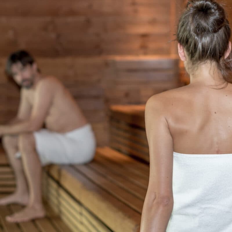 Zwei mit Handtüchern bekleidete Personen befinden sich in einer hölzernen Sauna; die eine sitzt im Hintergrund, während die andere mit dem Rücken zur Kamera im Vordergrund steht.