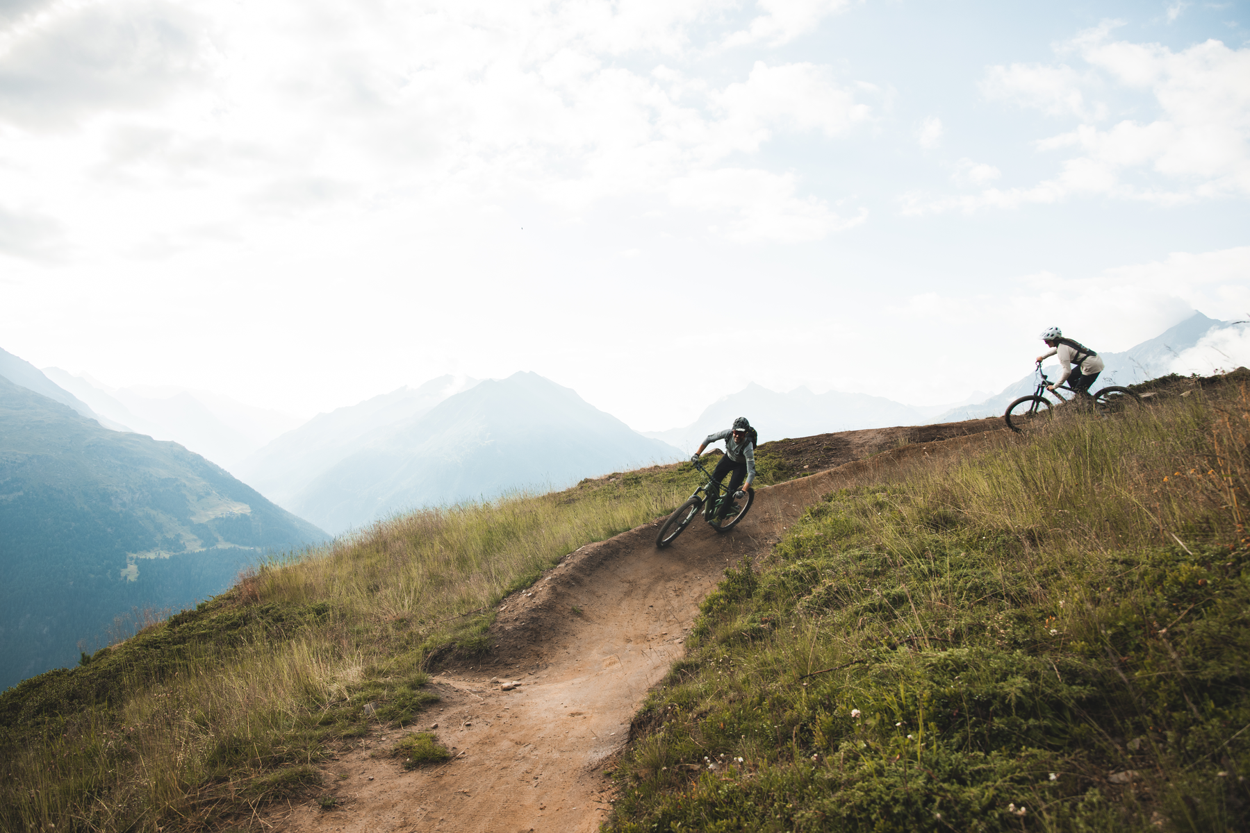 Two cyclists ride down a dirt trail on a grassy mountainside, surrounded by distant mountains under a partly cloudy sky.