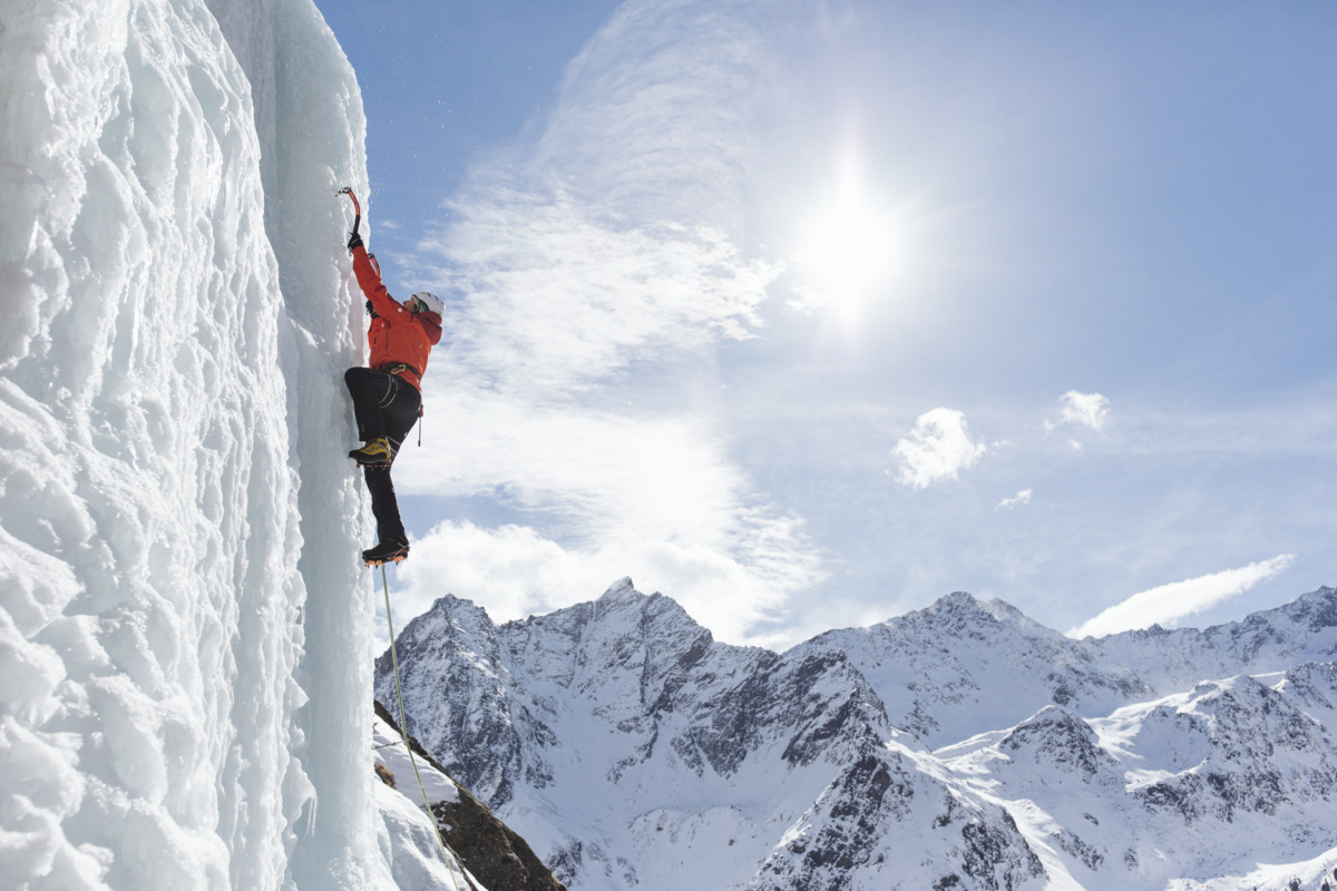 A person in an orange jacket climbs a steep ice wall with ice axes, with snowy mountains and a bright sun in the background.