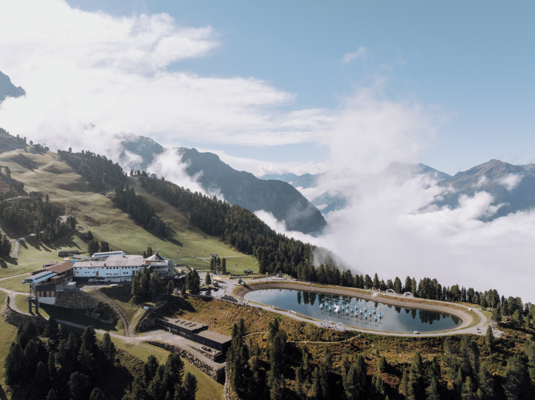 A mountain resort complex sits beside a circular pond, surrounded by trees and hills, with clouds and distant mountains in the background.