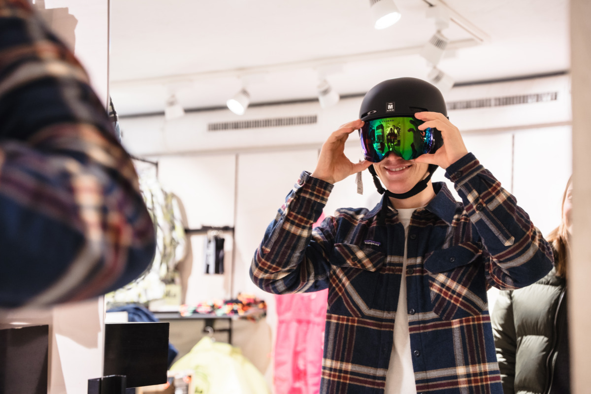 A person in a plaid shirt tries on a black ski helmet and reflective goggles in a store, standing in front of a mirror.