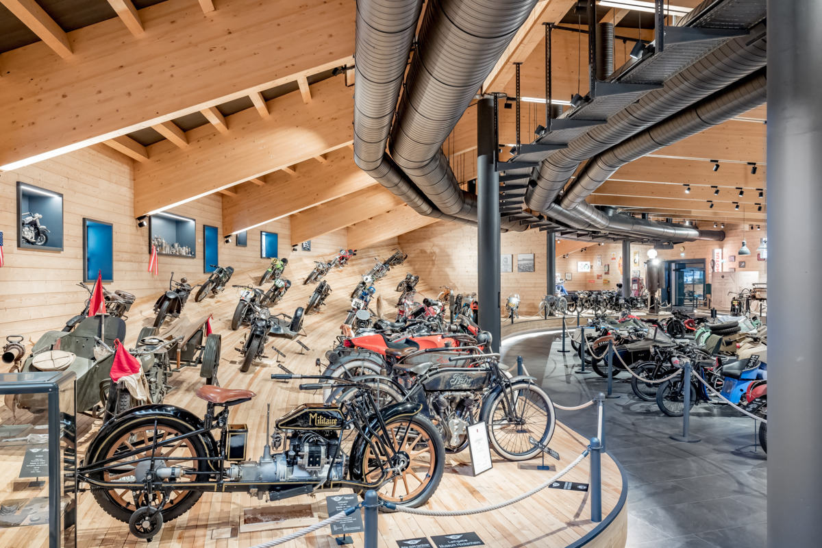 A spacious museum hall featuring a variety of vintage motorcycles displayed on wooden floors, with wood-paneled walls and exposed ceiling beams.