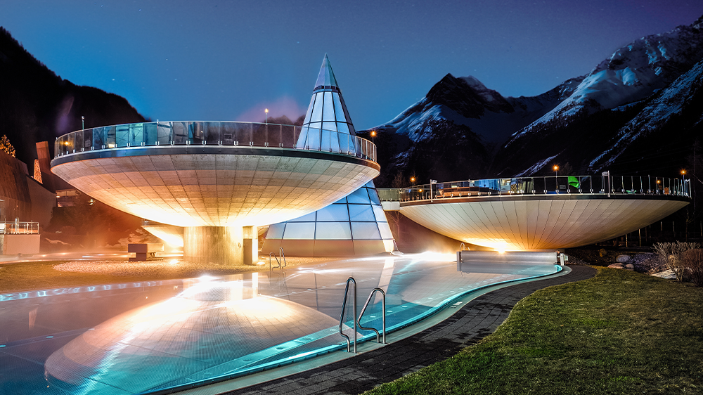 Futuristische, kuppelförmige Gebäude und eine kegelförmige Glasstruktur werden neben einem Außenpool beleuchtet, wobei in der Abenddämmerung die Berge im Hintergrund zu sehen sind.