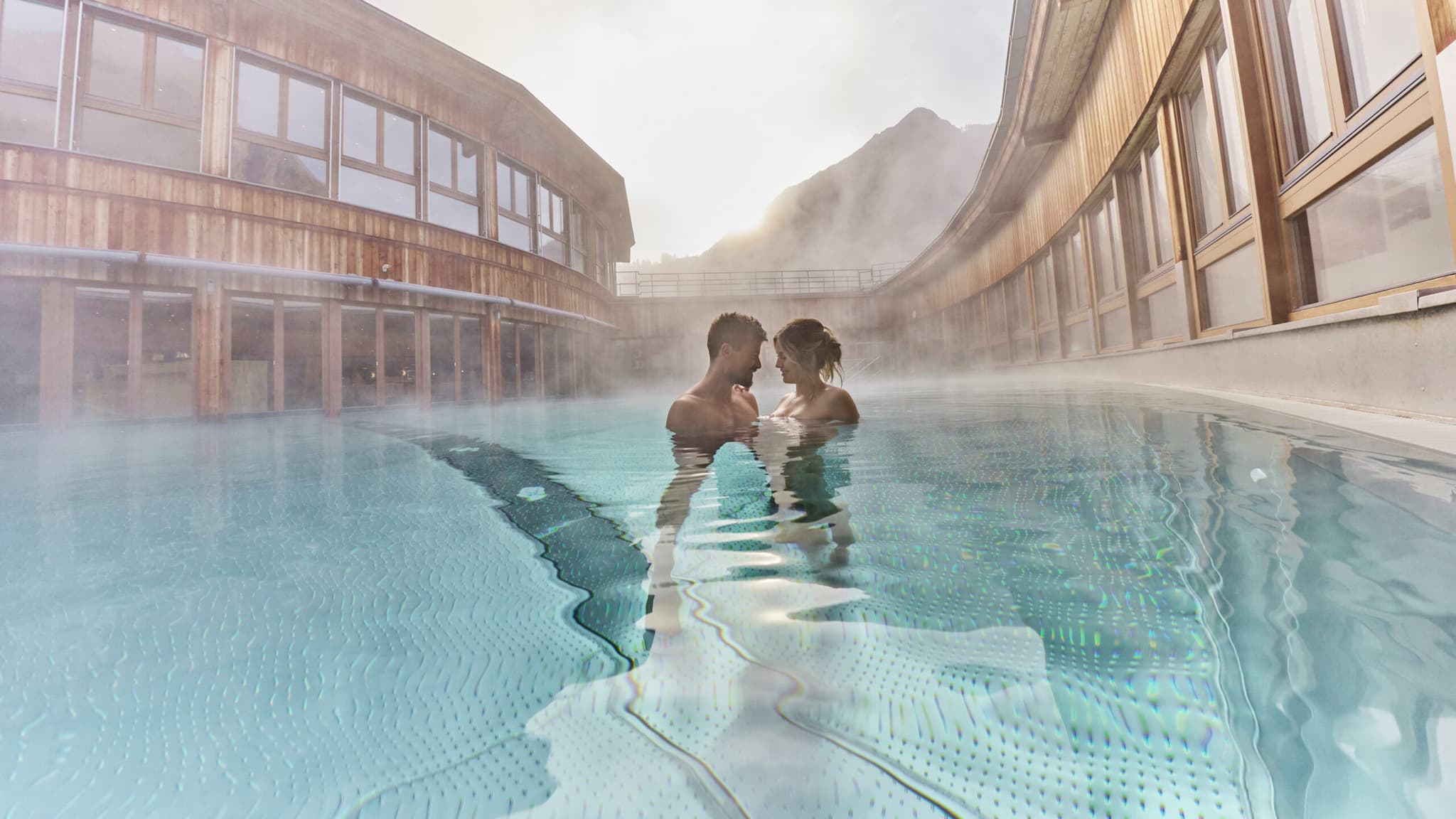 A couple stands close together in an outdoor pool surrounded by modern wooden buildings, with steam rising and mountains visible in the background.