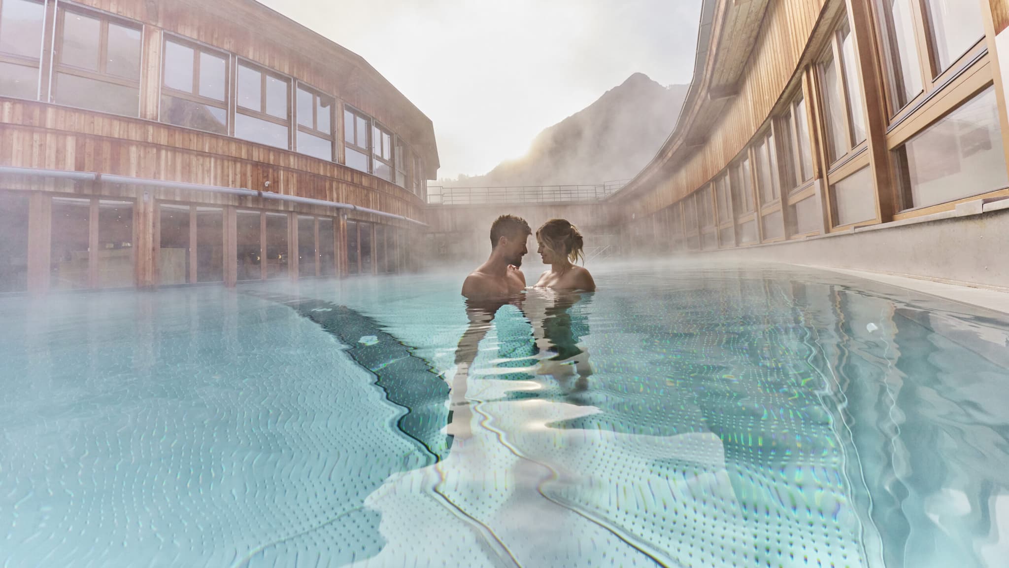 Due persone stanno una accanto all’altra in una piscina all’aperto riscaldata, circondata da moderni edifici in legno, con la nebbia che sale dall’acqua e le montagne visibili sullo sfondo.