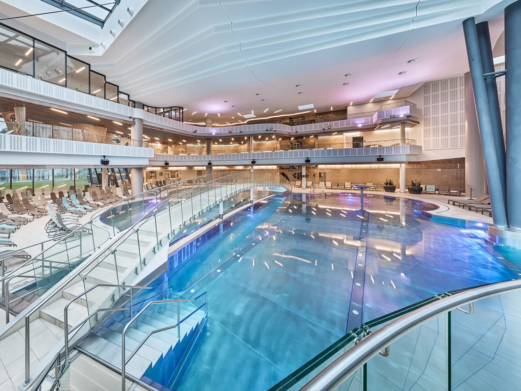 Indoor swimming pool with glass railings, lounge chairs, and multi-level balconies under bright modern lighting.