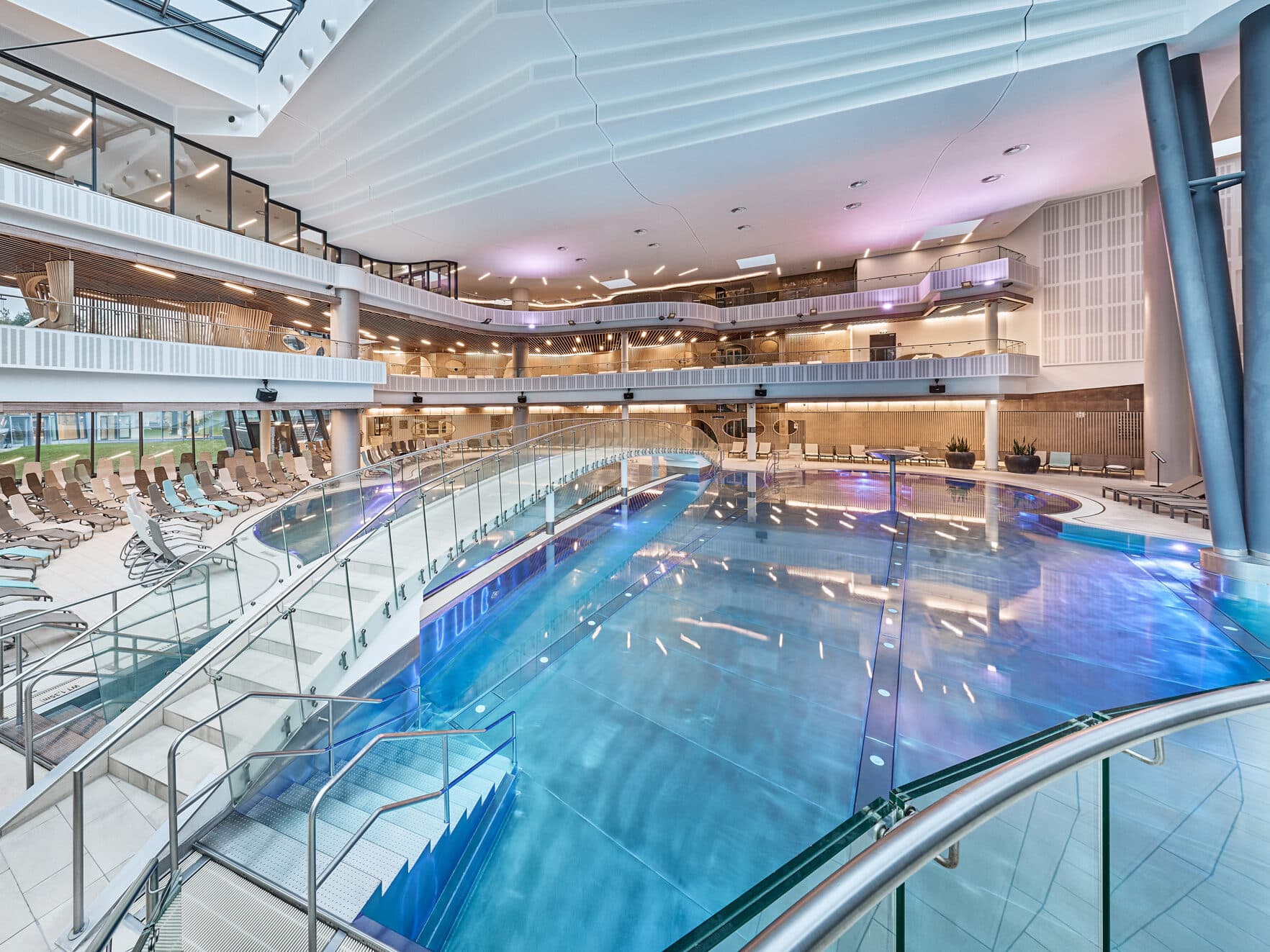 Piscine couverte aux eaux limpides, dotée de balustrades modernes en verre, de transats et de plusieurs niveaux, le tout baigné d’un éclairage artificiel éclatant.