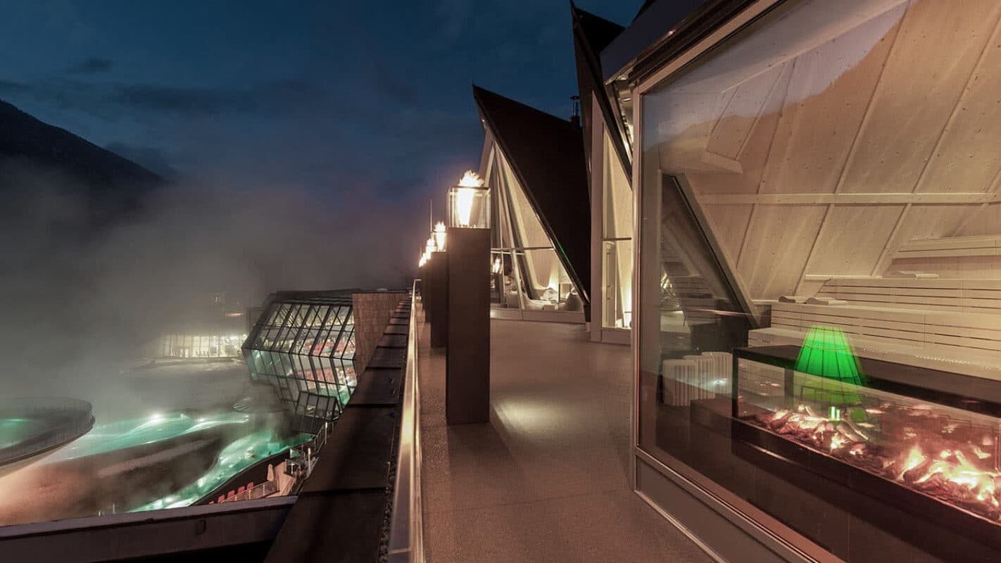 Modern building terrace at night with fire-lit lamps, large glass windows, interior seating, and outdoor pools with steam rising, set against a mountainous backdrop.