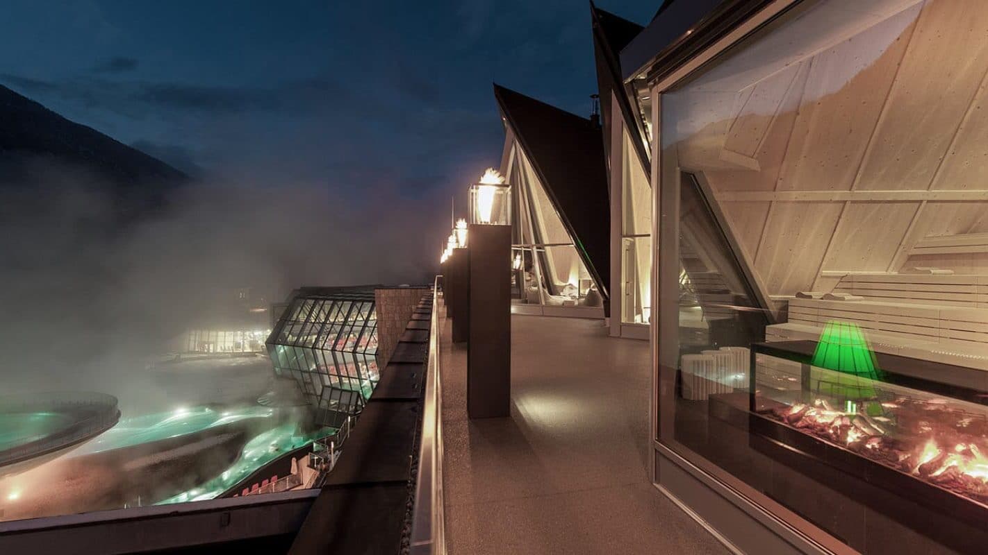 Terrazza di un edificio moderno di notte, con lampade accese, ampie finestre che riflettono le luci interne e, più in basso, un'area piscina all'aperto da cui sale il vapore e l'acqua verde è illuminata.