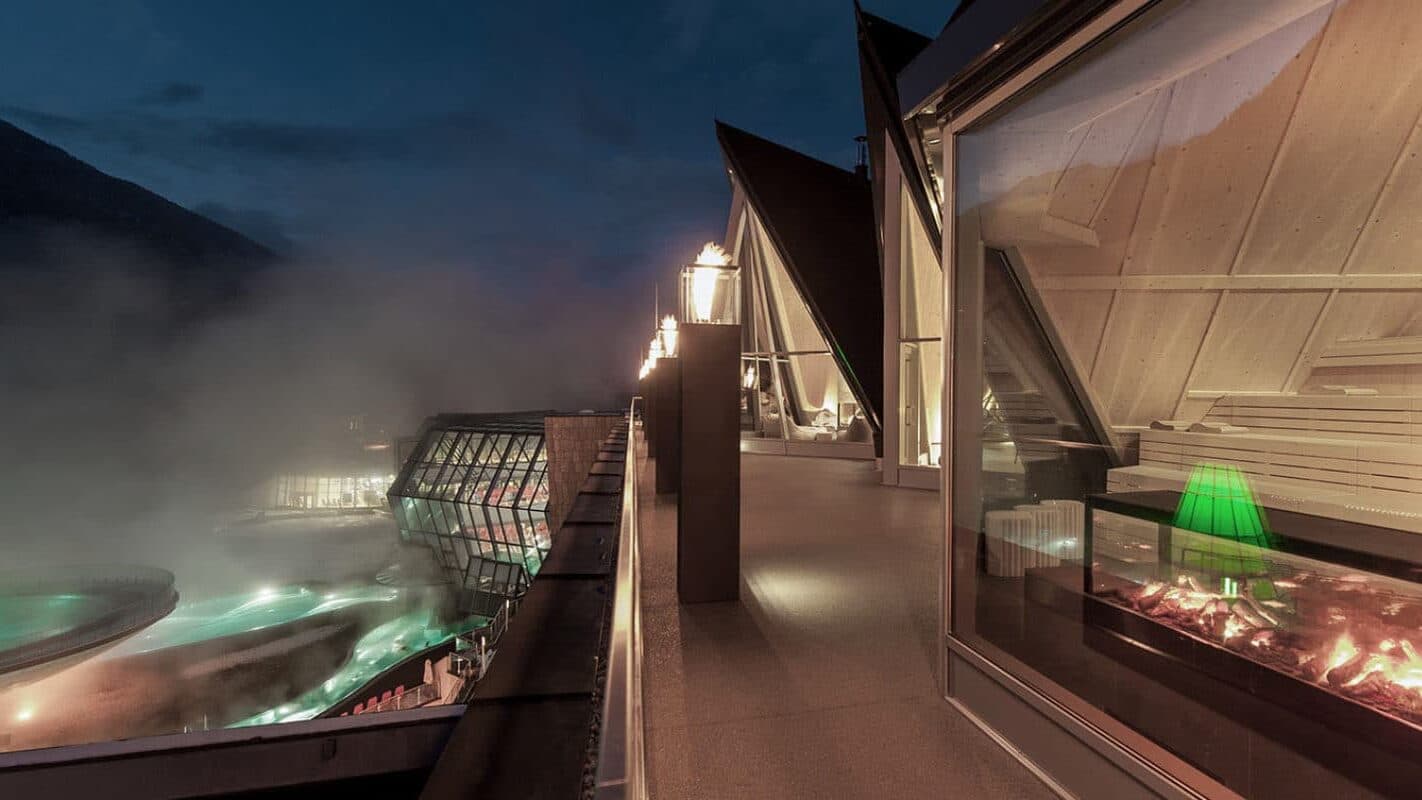 Un edificio moderno, con ampie vetrate e tetti spioventi, si affaccia sulle piscine all’aperto illuminate di notte, con la nebbia che sale sullo sfondo.