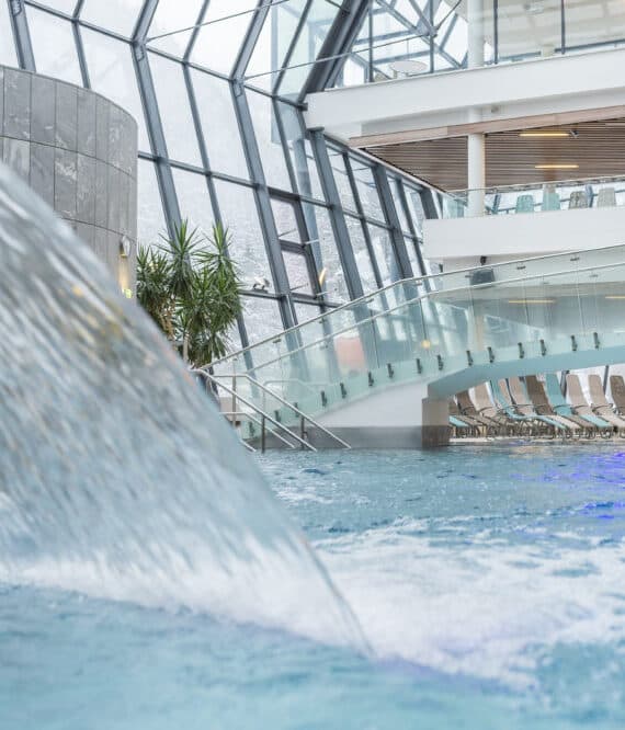 Piscina coperta con getti d'acqua, pareti in vetro e un ponte moderno. Accanto alla piscina si vedono sedie a sdraio e piante in vaso.