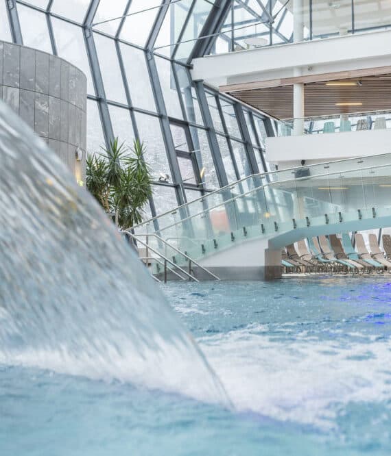 Piscina coperta con una parete di vetro curva, architettura moderna, una fontana ornamentale e sedie a sdraio sullo sfondo.
