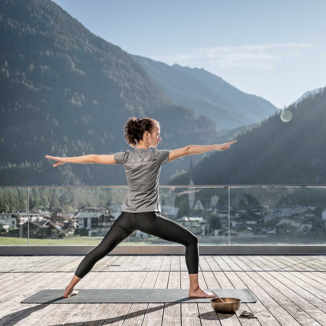 Una persona che pratica yoga nella posizione del Guerriero II su un tappetino all’aperto, con le montagne e un villaggio sullo sfondo in una giornata serena.