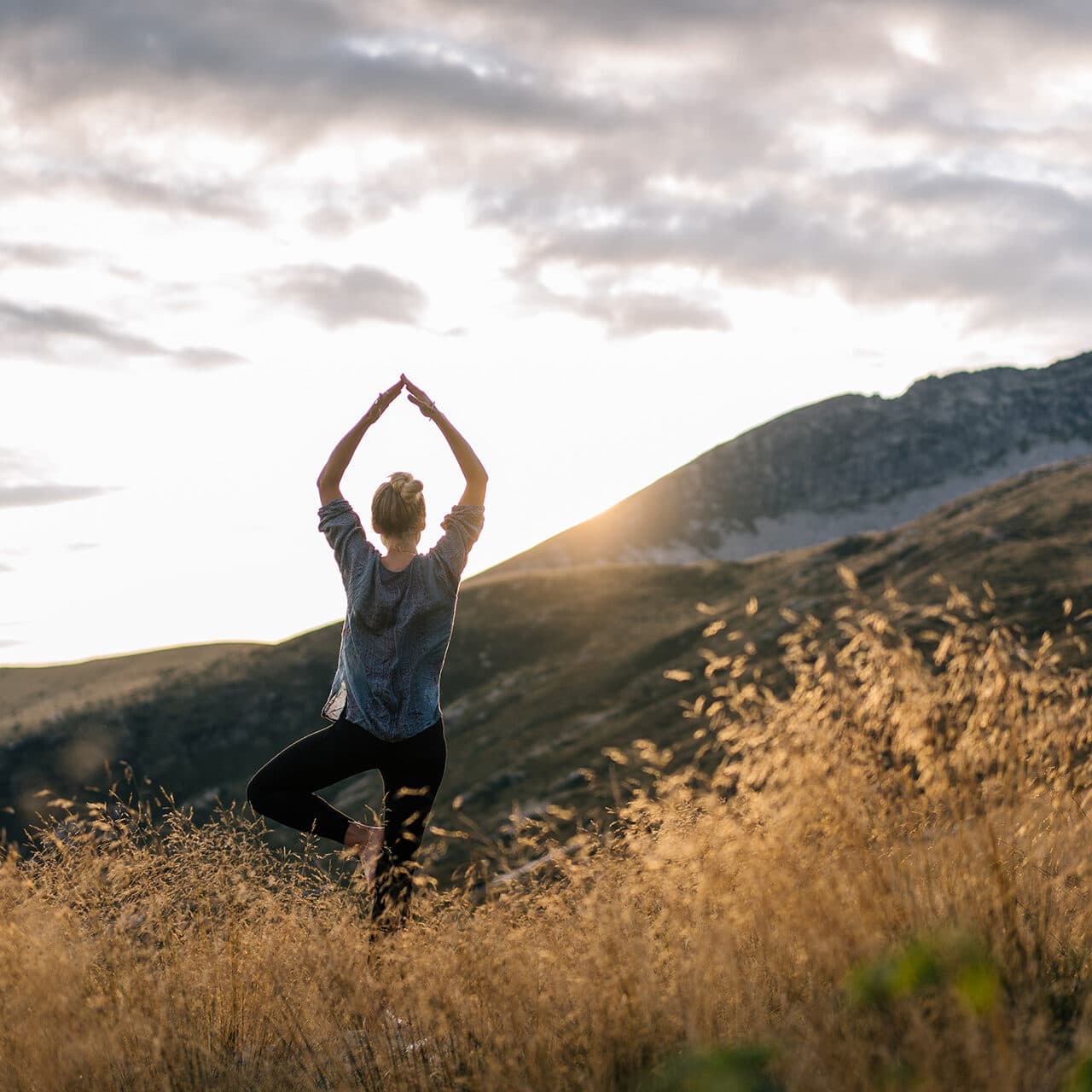 Person standing in a yoga pose on one leg in a grassy field at sunset, with mountains in the background.