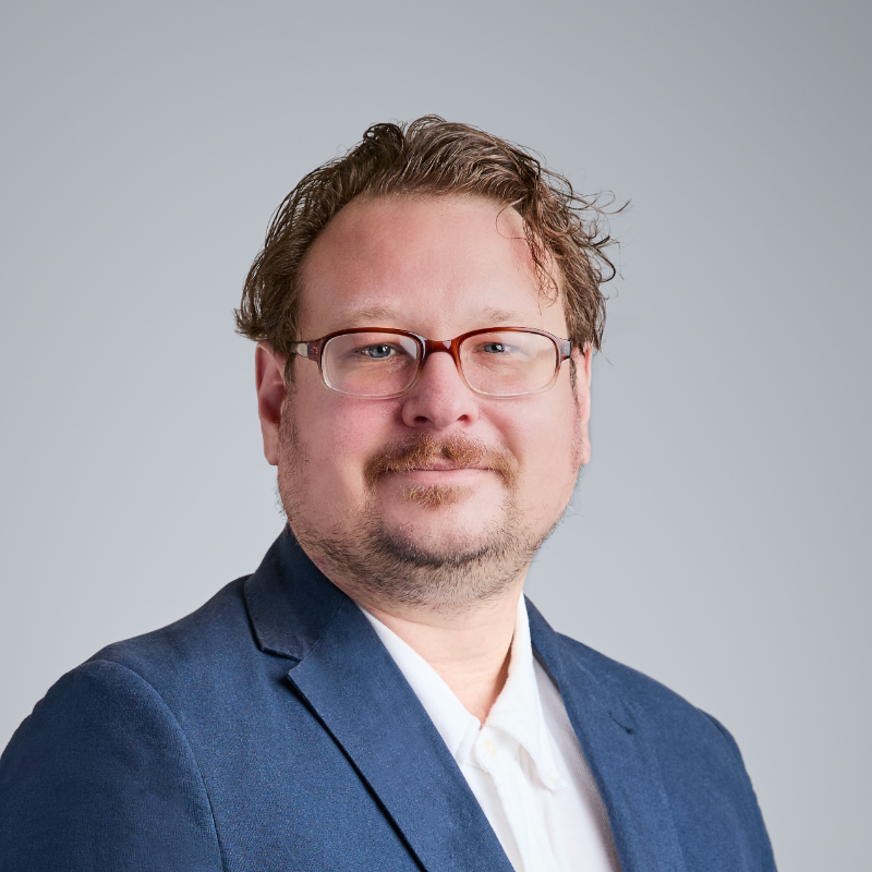 A middle-aged man with glasses, a goatee, and messy hair, wearing a white collared shirt under a blue blazer.
